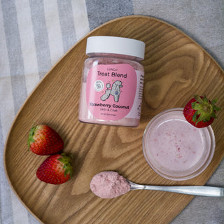 Jar of strawberry coconut treat blend on a wooden plate with strawberries and a spoonful of powder.