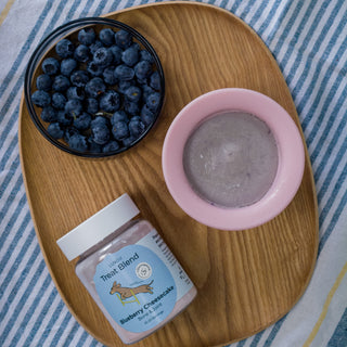 Wooden tray with blueberries, a pink bowl, and a jar of 'Teat Blend' on a striped fabric background.